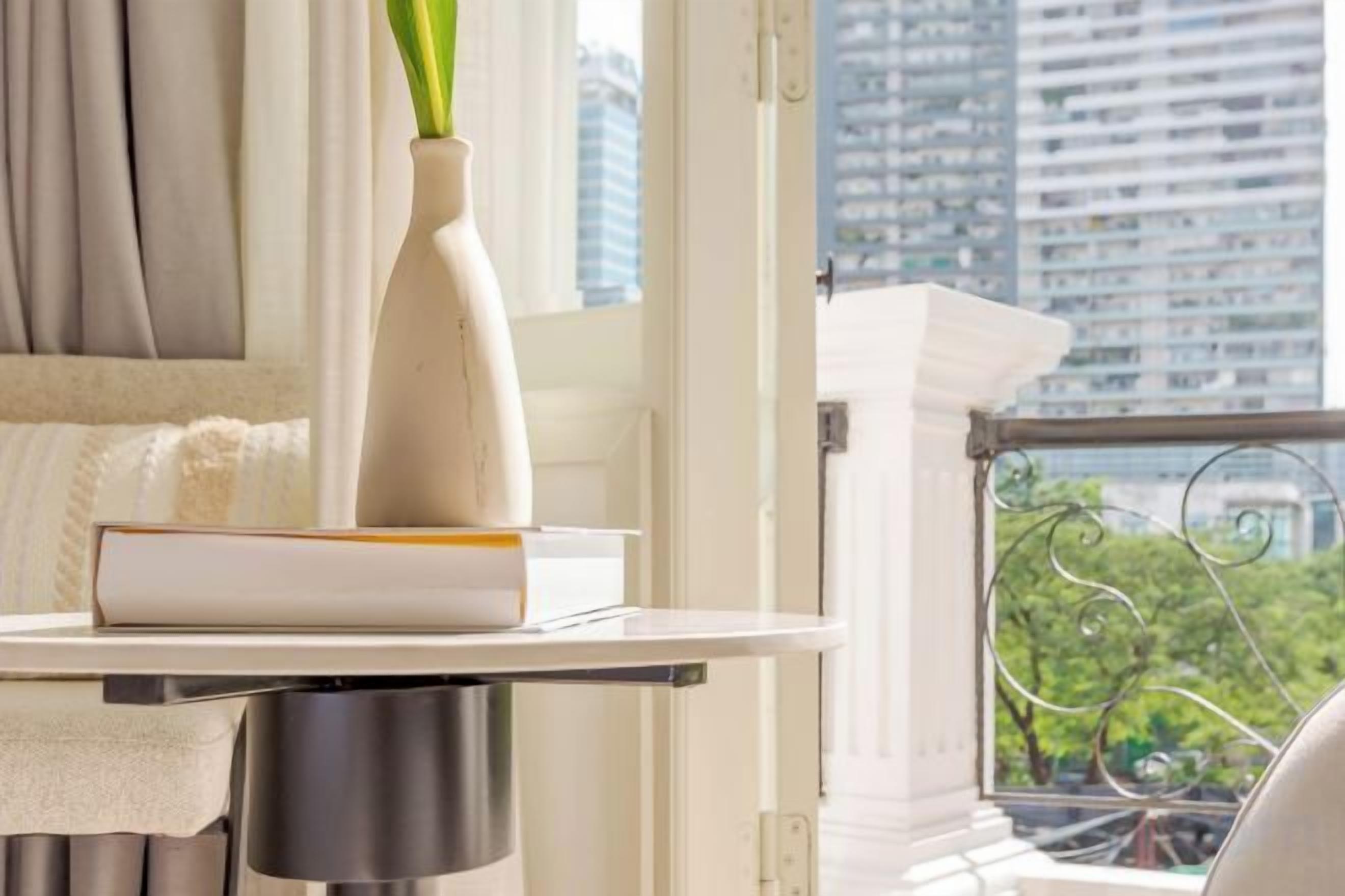 A table with a vase and a book, a window with a view of buildings, and a balcony railing.