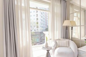 A room with a large window, a white chair, a small table with a vase, and a lamp. - Keypad Hotel - Nhân Hòa (Hanoi)