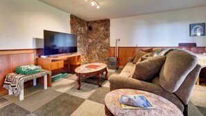 A living room with a stone accent wall, a flat-screen TV, a wooden entertainment unit, a brown sofa, and a patterned rug. - Fish & Hike! Home Base for Kachemak Bay Adventures (Homer)