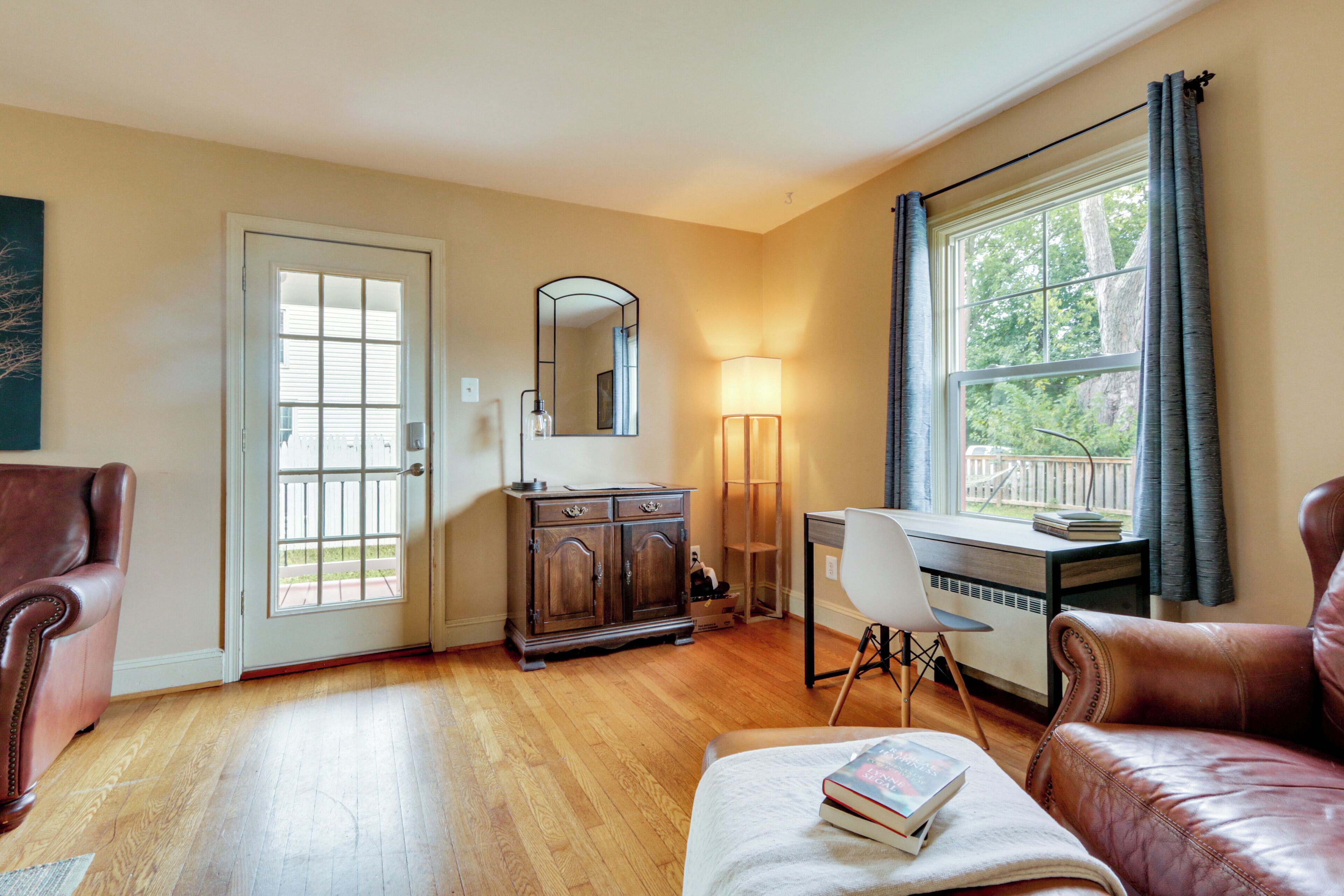 Ein Zimmer mit Holzboden, Schreibtisch mit Stuhl, Ledersessel, Schrank und ein großes Fenster mit Vorhängen.