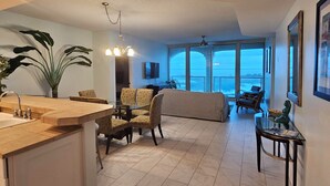 A spacious living area with a large window offering a view of the ocean, a dining area with a kitchen counter, and a seating area with patterned chairs.