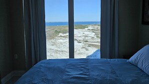 A bedroom with a large window offering a view of a beach and ocean.