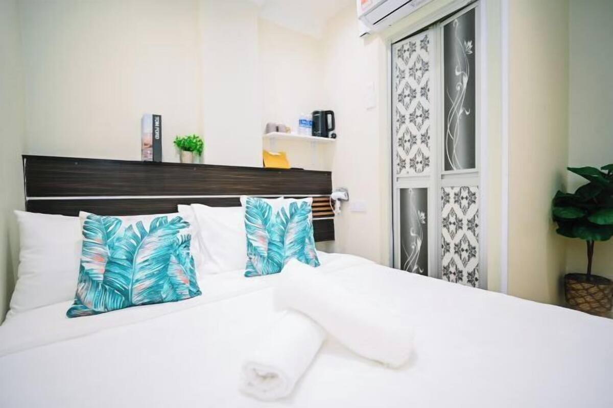 A neatly made bed with white bedding and tropical leaf-patterned pillows, a small shelf with toiletries, and a decorative plant beside the window.