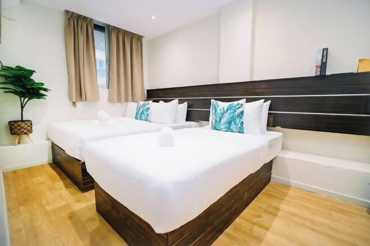 A hotel room with two single beds, a large window with curtains, a potted plant, and a wooden floor.