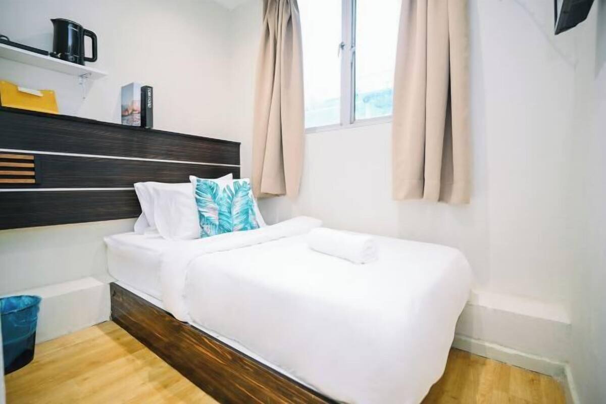 A small, neatly arranged bedroom with a single bed, a wooden headboard, a shelf with a kettle, and a window with curtains.