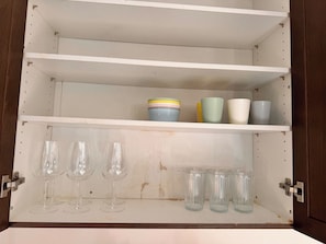 A cabinet with three shelves; the top shelf holds colorful bowls, the middle shelf has clear glass cups, and the bottom shelf contains wine glasses and more clear glass cups. - Modern Brampton Home | 3 BR 2.5 WR | Free Parking (Brampton)