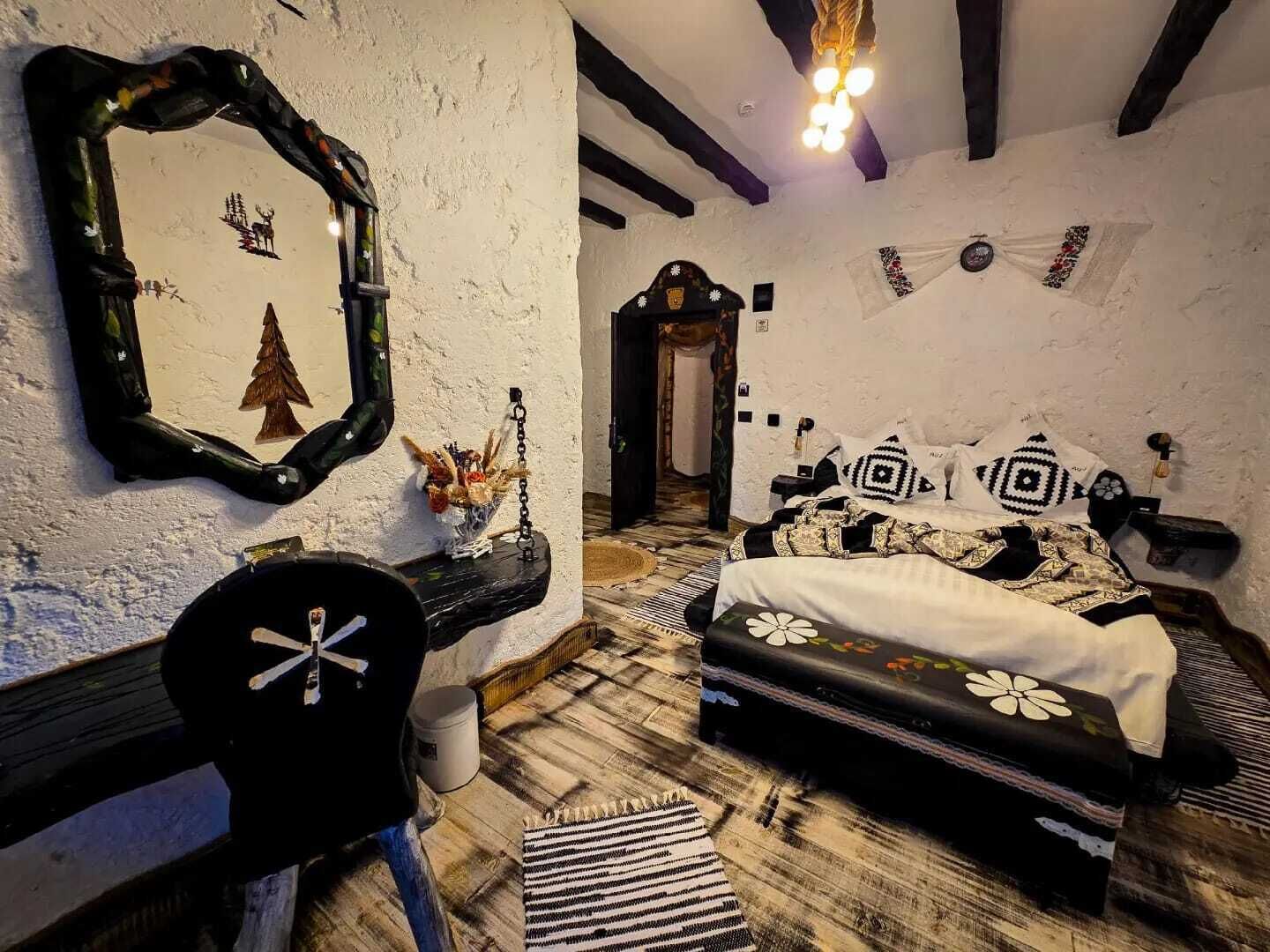 A rustic bedroom with a wooden floor, a bed with patterned bedding, a mirror with a tree design, and a small table with a vase.