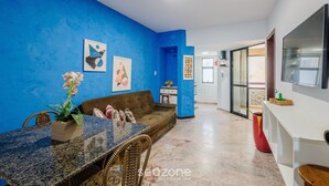 A living room with a blue wall, a brown sofa, a dining table with chairs, and a TV on the wall. - Fully Equipped Apartments Just Steps From Barra Beach and Barra Lighthouse - Csfs (Salvador)