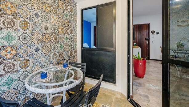 A dining area with a round table and chairs, a tiled wall with intricate patterns, and a sliding glass door leading to an outdoor space with a potted plant. - Fully Equipped Apartments Just Steps From Barra Beach and Barra Lighthouse - Csfs (Salvador)