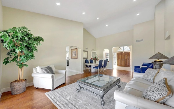 A spacious living room with a large plant, white furniture, and a dining area with blue chairs.