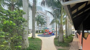 A pathway leading to a playground with a slide and climbing structure, surrounded by palm trees and a building with balconies.
