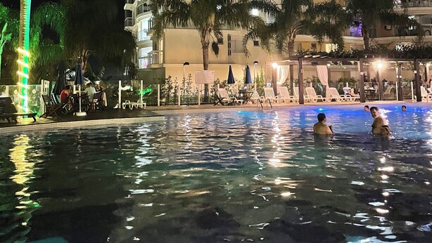 A nighttime view of a hotel with a swimming pool, palm trees, and outdoor seating areas.