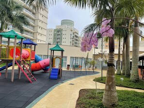 Ein Spielplatz mit Rutschen und Klettergeräten, umgeben von Palmen und einem Wohnhaus im Hintergrund.