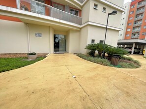 Entrada de um edifício com um caminho de concreto que leva a uma porta de vidro, cercada por vegetação e uma lixeira.