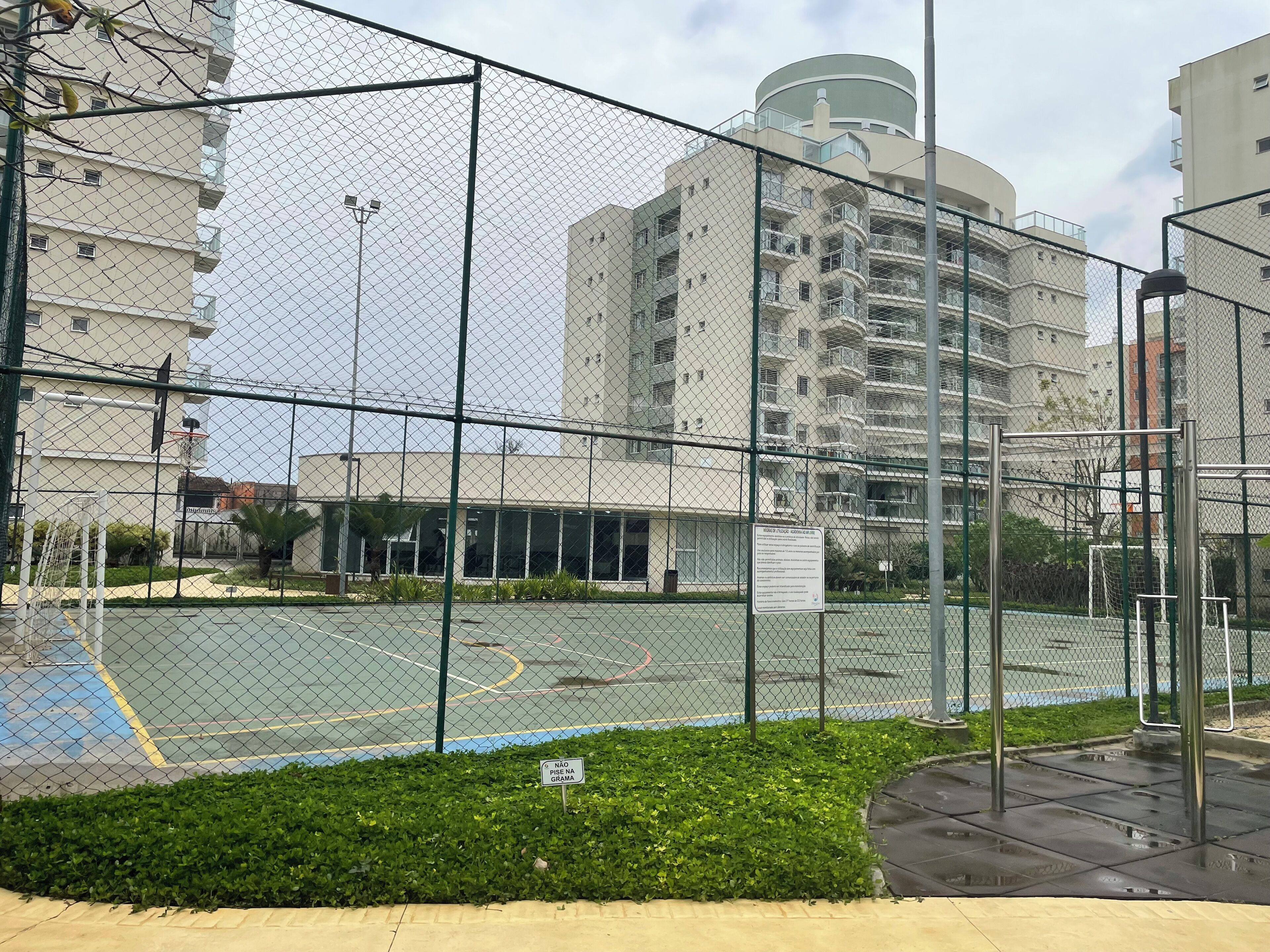 Un terrain de sport délimité par une clôture, avec un panier de basket, le tout entouré d’une clôture en treillis métallique.
