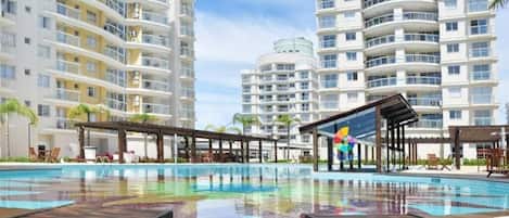 A swimming pool with wooden loungers, surrounded by multi-story buildings with balconies.