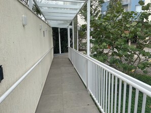 Um corredor externo coberto, com guarda-corrimão branco, piso de concreto e vista para um edifício e vegetação ao fundo.
