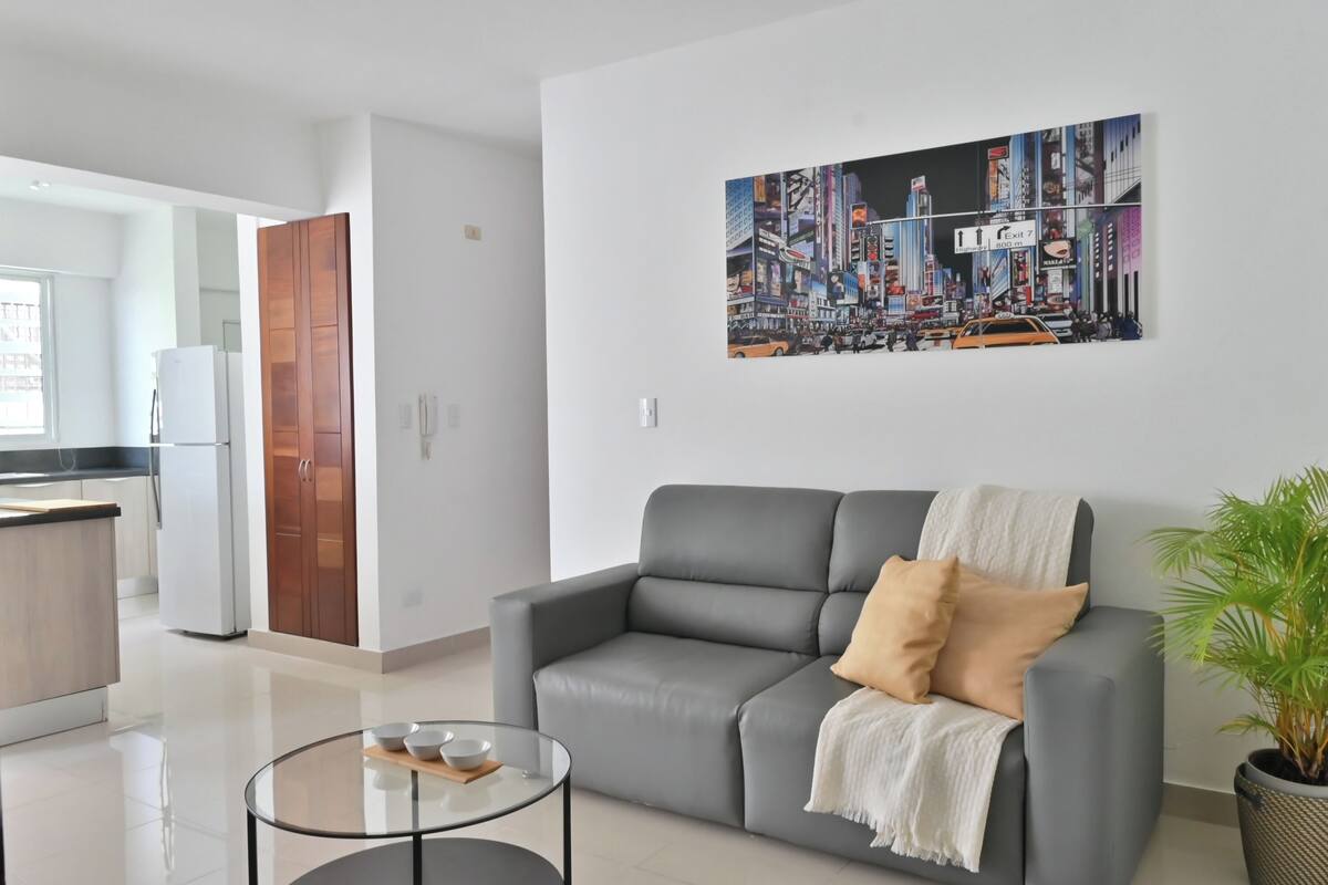 Una sala de estar moderna con un sofá gris, una mesa de centro de vidrio y una pintura de un paisaje urbano en la pared.
