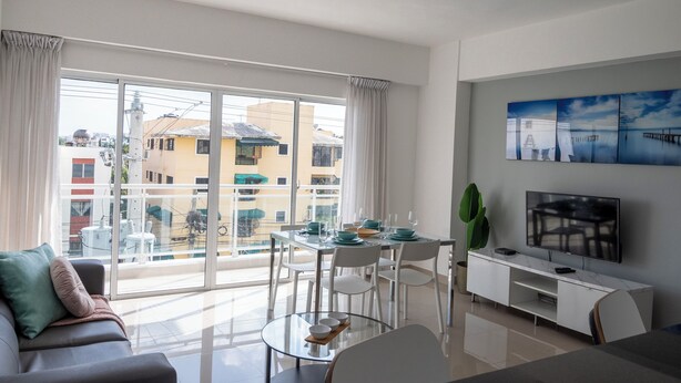 Un salon moderne avec une table à manger en verre, des chaises blanches, un canapé et un téléviseur posé sur un meuble blanc.