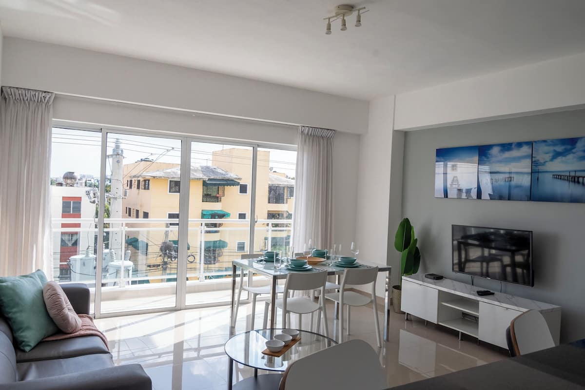 Una sala de estar moderna con una mesa de comedor de vidrio, sillas blancas, un sofá y un televisor sobre un mueble blanco.