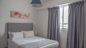 Une chambre à coucher comprenant un lit, une tête de lit, une table de chevet, une lampe, une fenêtre avec des rideaux et une œuvre d’art accrochée au mur.