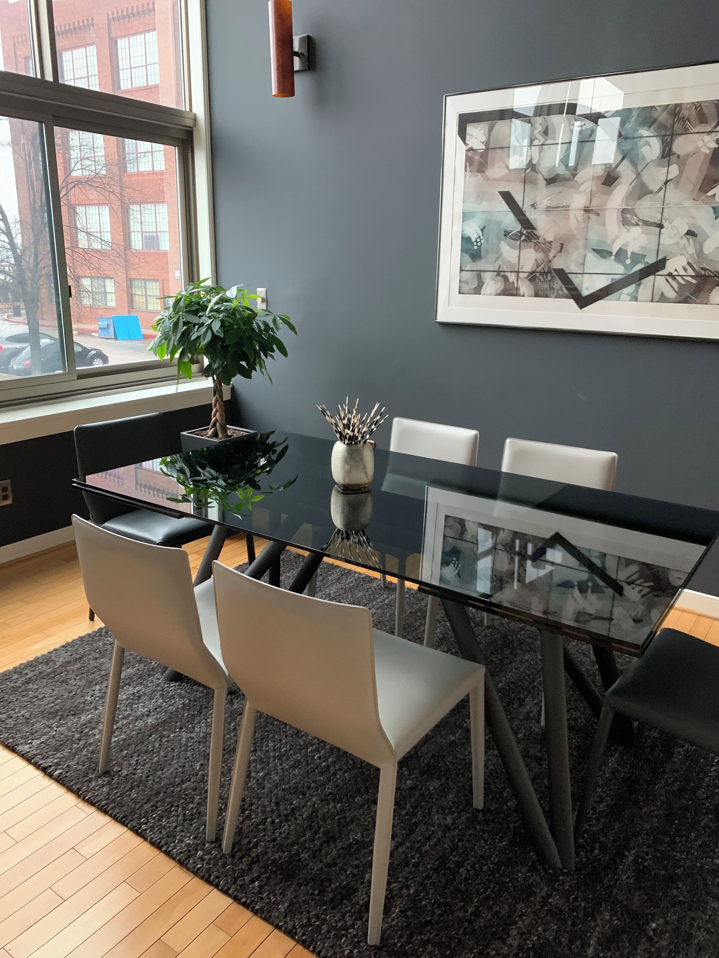 Un espace repas moderne avec une table en verre, des chaises blanches, une plante en pot et un tableau abstrait encadré accroché au mur.