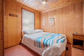 Una habitación con paneles de madera, una cama, una mesita de noche y un ventanal con persianas.
