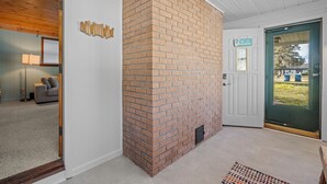 A brick fireplace in a room with white walls and a wooden ceiling. A door with a green frame and a sign above it is visible. - Quaint 2-bedroom cottage in Houghton Lake - quick walk to lake (Houghton Lake)