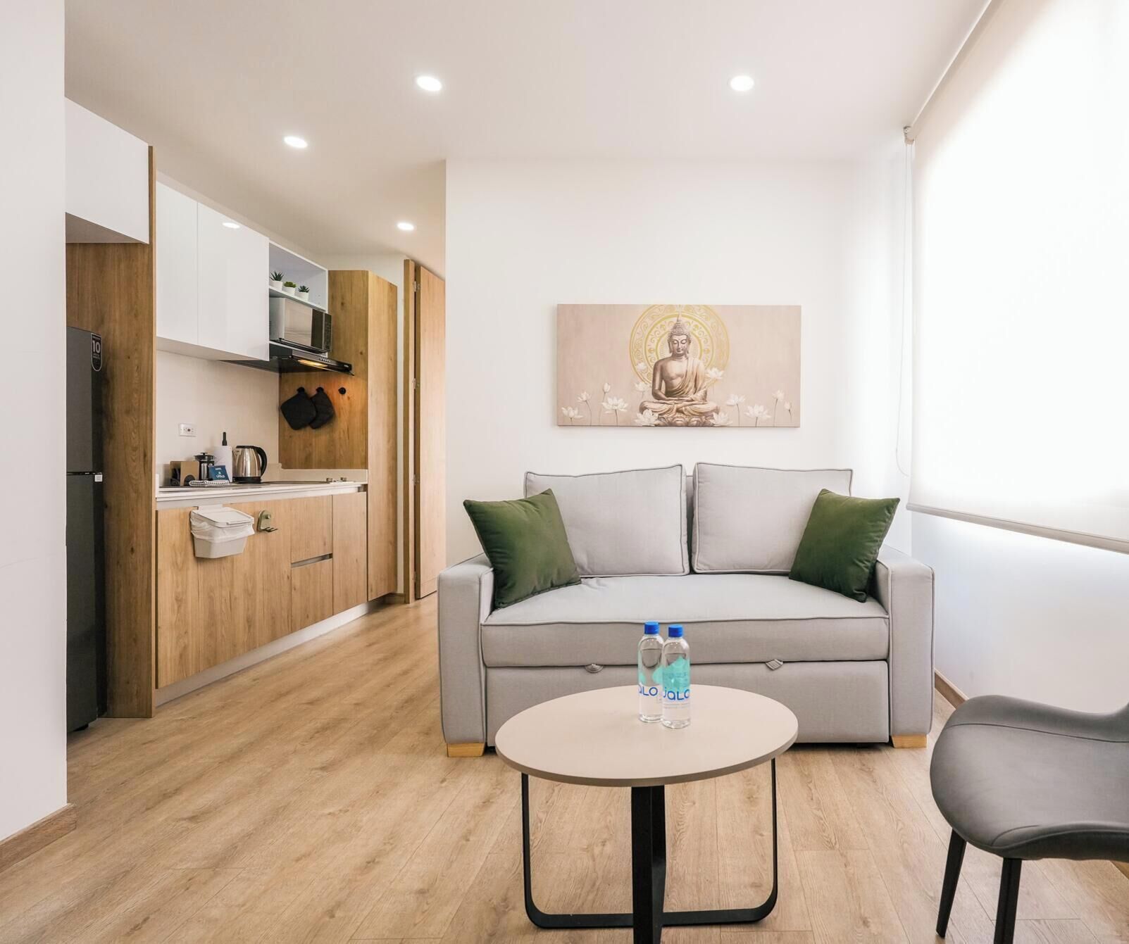 Uma sala de estar moderna com um sofá cinzento, uma mesa de centro redonda e uma área de cozinha com armários de madeira e uma pintura de Buda.