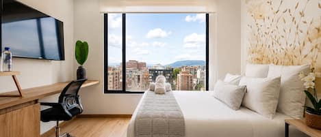 A modern bedroom with a large bed, a desk, a chair, a TV, and a window with a city view.