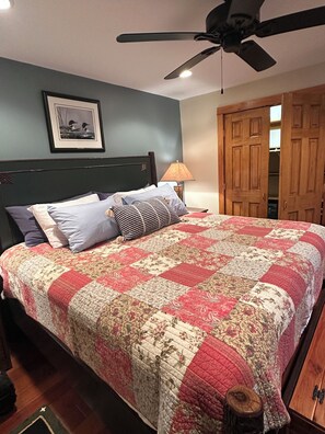 A bedroom with a large bed, a ceiling fan, a wooden wardrobe, and a framed picture on the wall.