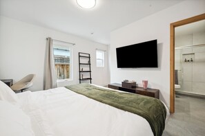A hotel room with a bed, a TV mounted on the wall, a desk with a chair, a window with curtains, and a bathroom visible through an open door. - NEW, fast WI-FI, in unit W/D (San Jose)