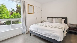 Une chambre à coucher avec un lit, une table de chevet, une fenêtre donnant sur un espace verdoyant et une porte.