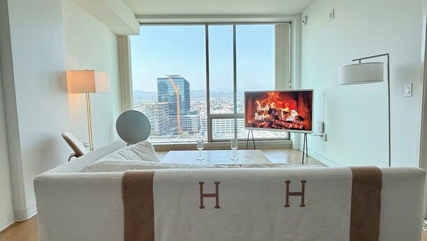A hotel room with a large window, a flat-screen TV, a bed with a 'HH' headboard, and a city view. - Ultra Modern 1BD 1BA Flats in heart of LA - true gem (Los Angeles)