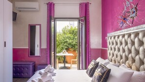 A hotel room with a bed, a table with chairs, a purple sofa, and a view of greenery outside.