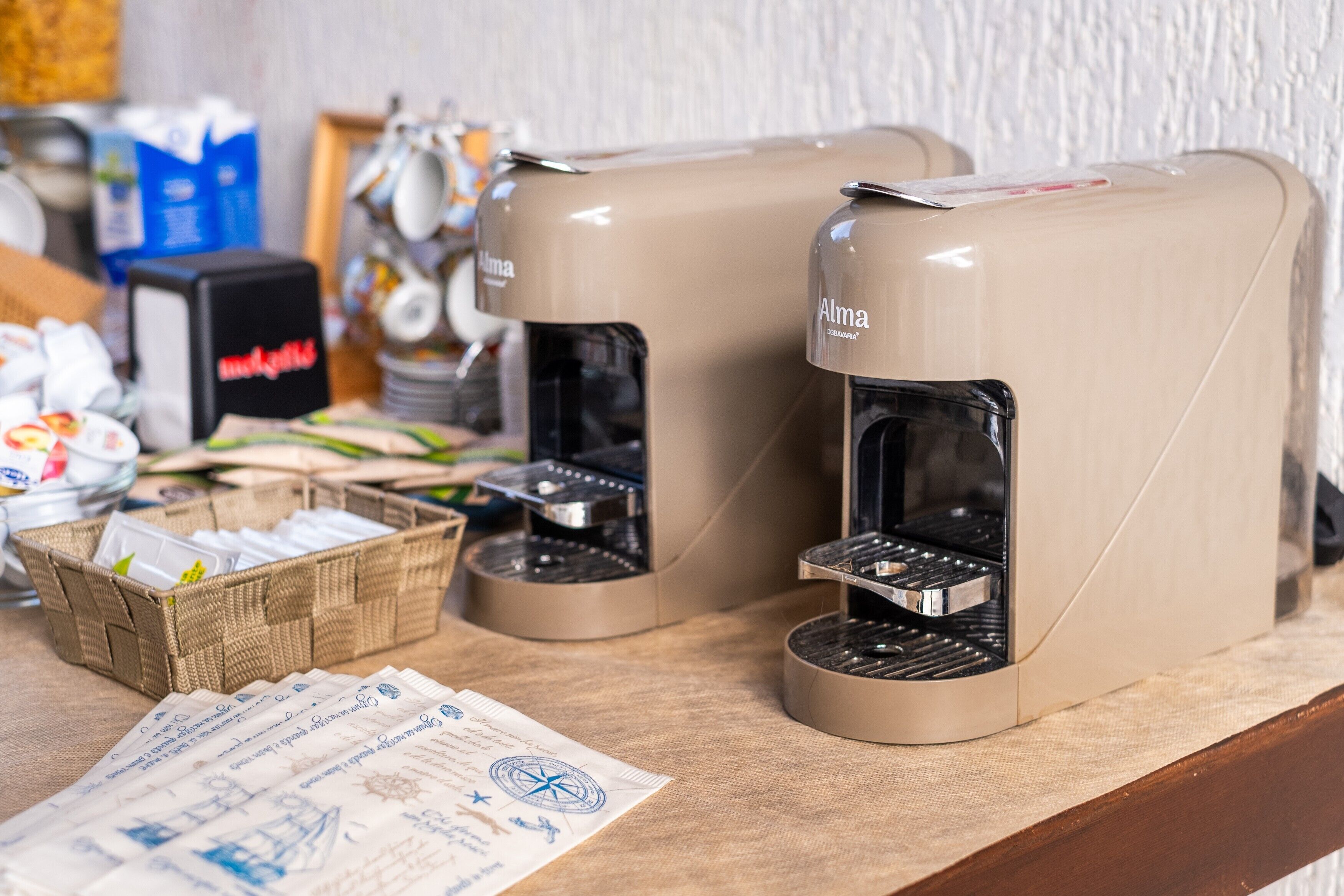 Two coffee machines with the brand 'Alma' on a counter with a wicker basket and a menu.
