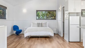 A bedroom with a bed, a window, a blue chair, and a kitchen area with a refrigerator.