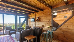 Un interior de madera con un sofá, sillas, una mesita, televisión y vistas a través de amplias ventanas.