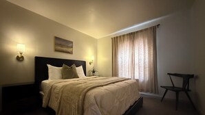 A neatly made bed with a headboard, two bedside lamps, a painting on the wall, and a chair in the corner. - Cozy  Coastal by Beaches & Redwoods (McKinleyville)
