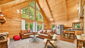 A spacious wooden cabin with a large living area featuring a leather sofa, a coffee table, and a fireplace. The kitchen area is visible with a refrigerator and a sink. - Log Cabin Luxury Near IU & Lake Monroe (Bloomington)