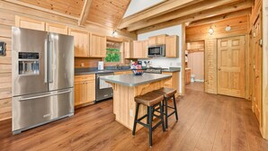 A modern kitchen with wooden cabinets, a stainless steel refrigerator, a microwave, and a large island with stools. - Log Cabin Luxury Near IU & Lake Monroe (Bloomington)
