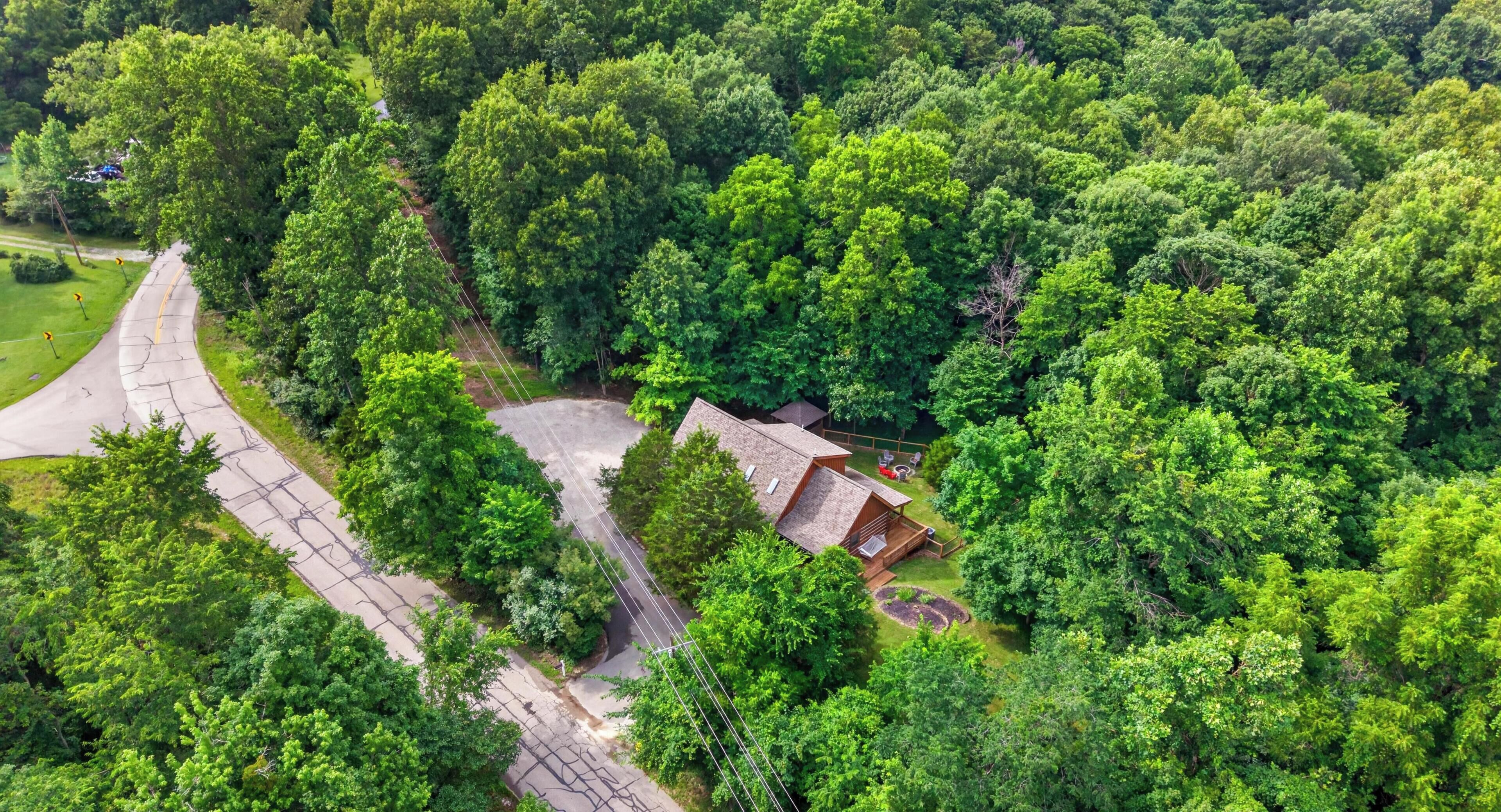 An aerial view of a property surrounded by dense greenery, featuring a winding road, a small building with a deck, and a clear sky.