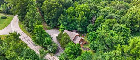 An aerial view of a property surrounded by dense greenery, featuring a winding road, a small building with a deck, and a clear sky.