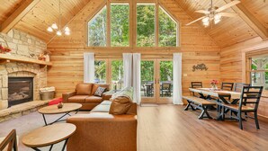 A cozy cabin interior with a stone fireplace, wooden walls, a large dining table, and a ceiling fan. - Log Cabin Luxury Near IU & Lake Monroe (Bloomington)