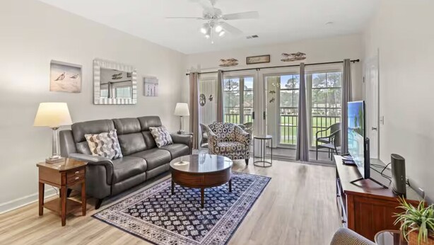 A modern living room with a leather sofa, a coffee table, and a patterned rug. There is a television, a desk with a computer, and a chair. The room has a ceiling fan and large windows with curtains. - Sandy Knees Retreat - Steps to Golf and Minutes to Sunset Beach (Sunset Beach)