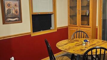 Un coin repas comprenant une table ronde, quatre chaises, une fenêtre avec des stores, un cadre photo fixé au mur et un buffet en bois avec des portes en verre.