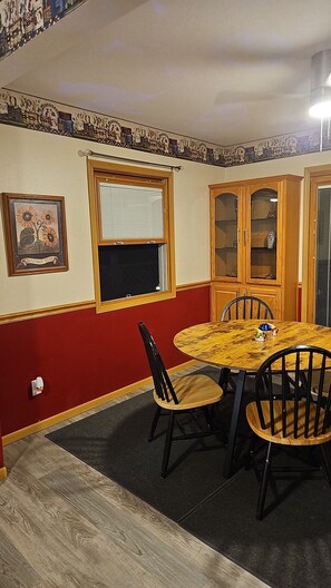 A dining area with a round table, four chairs, a window with blinds, a wall-mounted picture frame, and a wooden cabinet with glass doors.