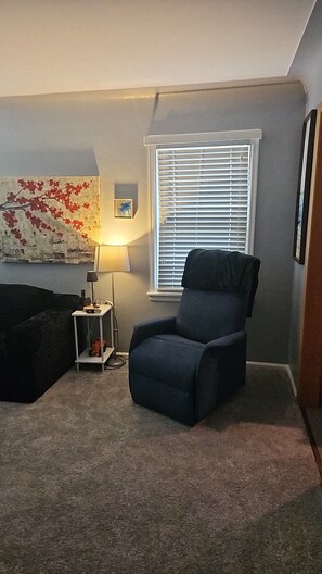 A living room with a dark blue armchair, a lamp, a small table with a lamp, a painting on the wall, and a window with blinds.