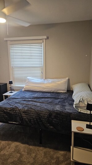 A bedroom with a bed, a bedside table, a lamp, a window with blinds, and a ceiling fan.