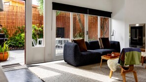 A modern living room with a sofa, a coffee table, and a TV. There is a sliding glass door leading to an outdoor area with a bamboo fence and plants.
